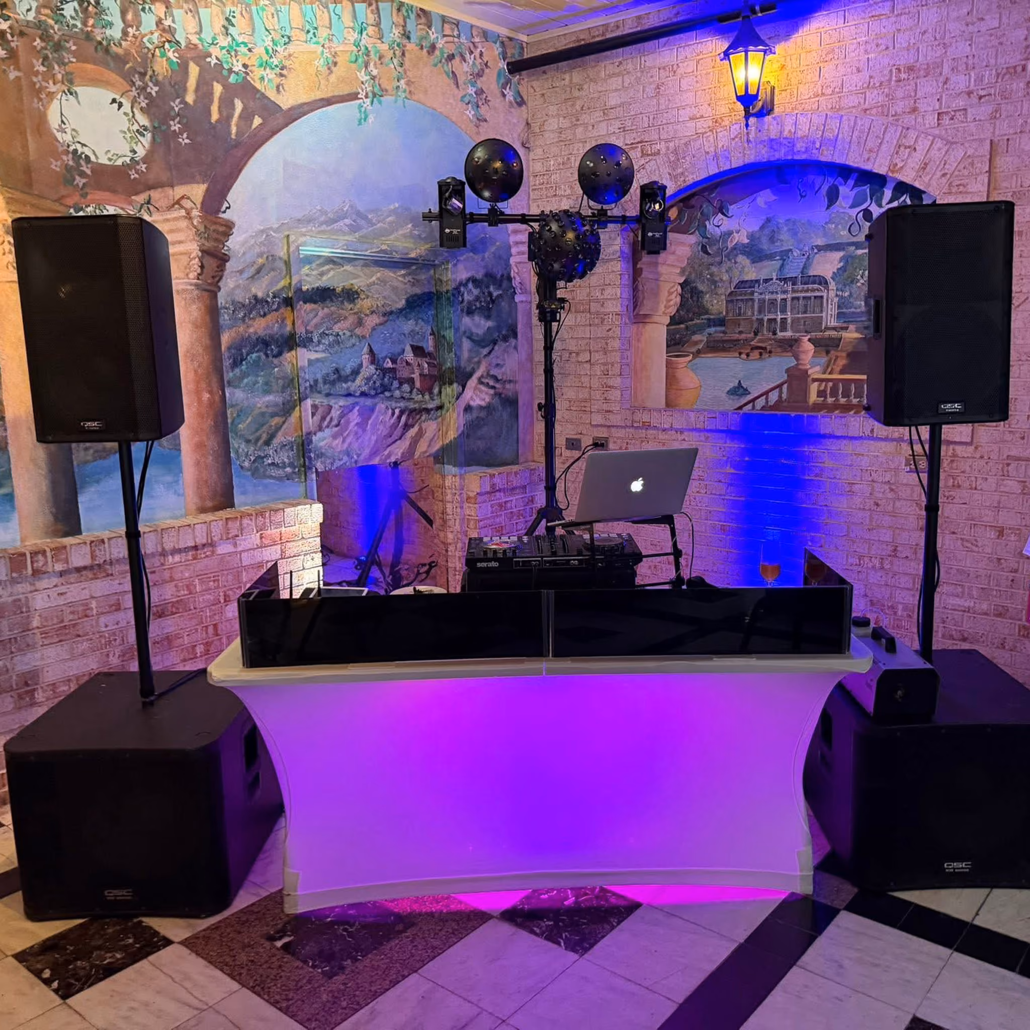 DJ booth with purple LED uplighting, Serato controller, laptop, moving head lights, and QSC speakers on stands, set against a painted Tuscan-style mural backdrop.