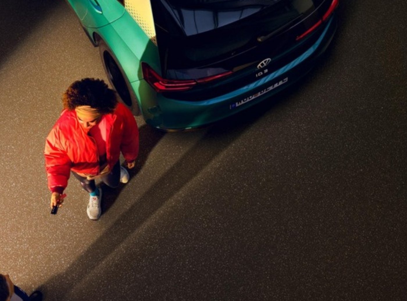 Uma : vue d’en haut, une femme en doudoune rouge près de l’arrière d’une voiture turquoise, sur un parking