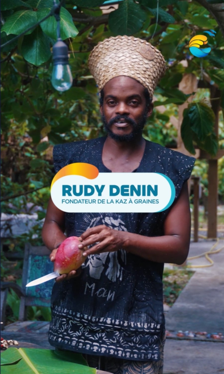 Office de Tourisme de la Riviera du Levant, « Les Visages du Levant » : le fondateur de la Kaz à Graines, coiffé d’un chapeau tressé, une mangue et un couteau à la main, dans son jardin