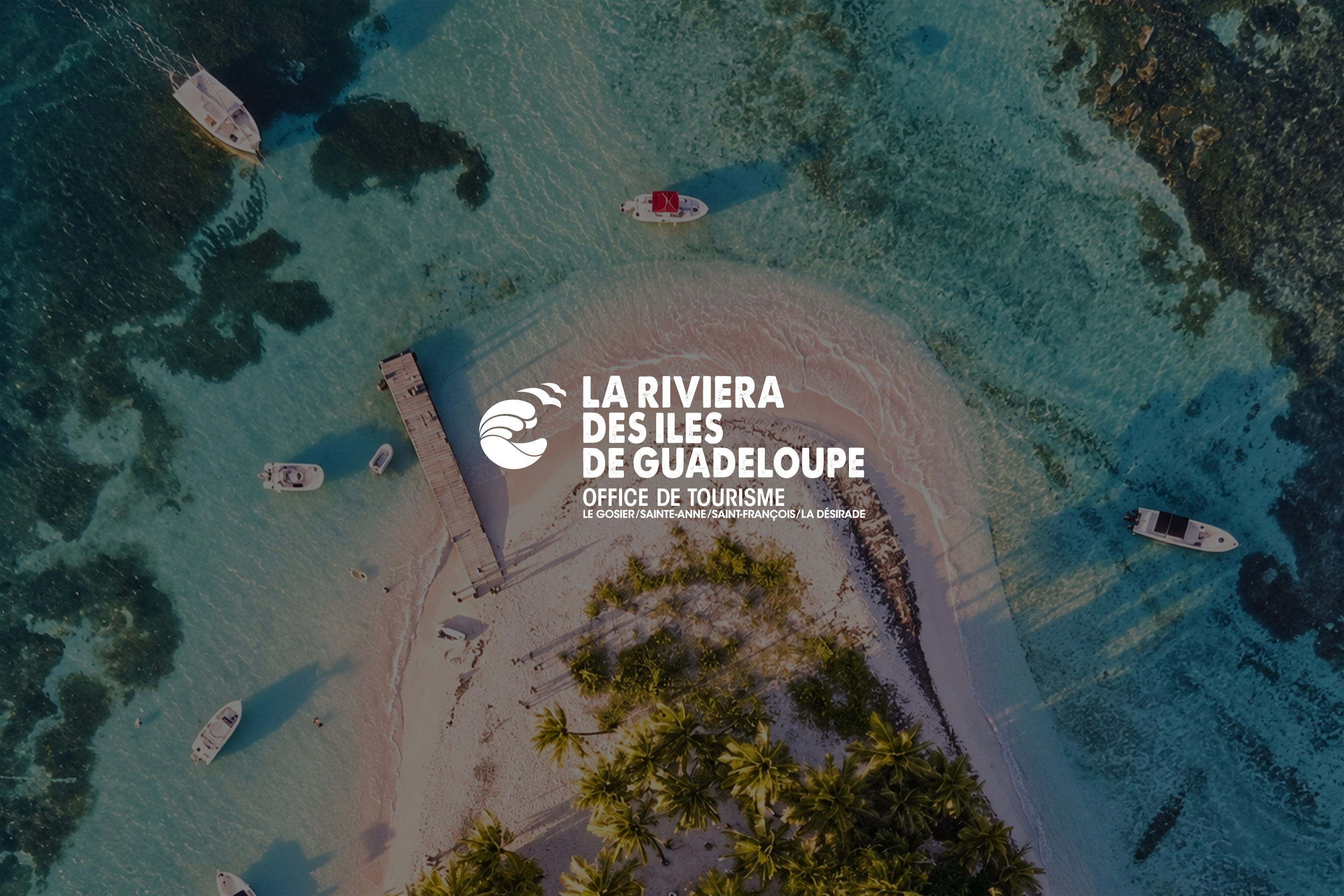 Office de Tourisme de la Riviera du Levant : le logo « La Riviera des Îles de Guadeloupe » sur la vue aérienne d’un îlet, de son ponton et de bateaux dans le lagon