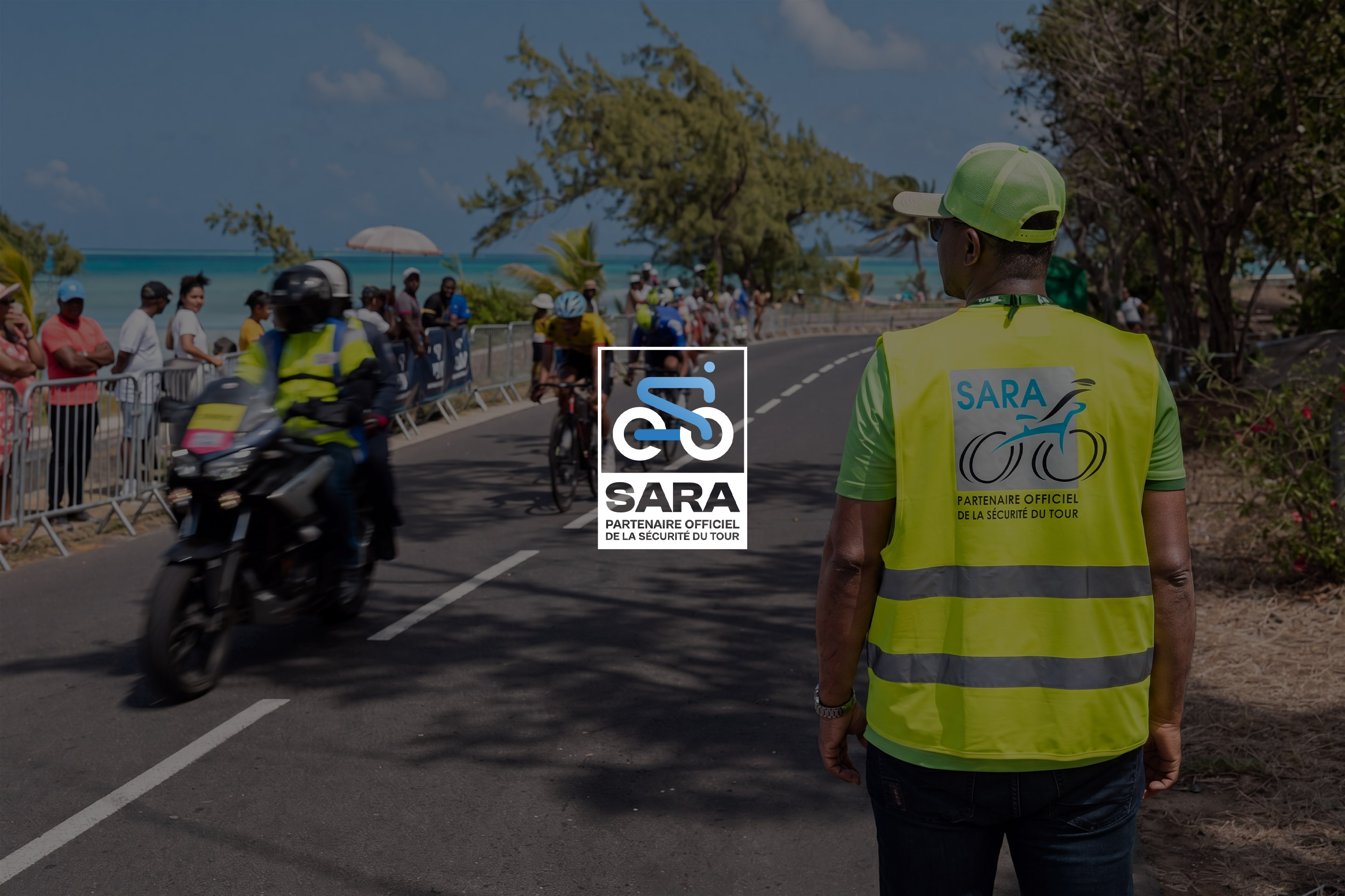 SARA, partenaire officiel de la sécurité du Tour : de dos, un signaleur en gilet jaune regarde passer une moto et un coureur, le long de la mer