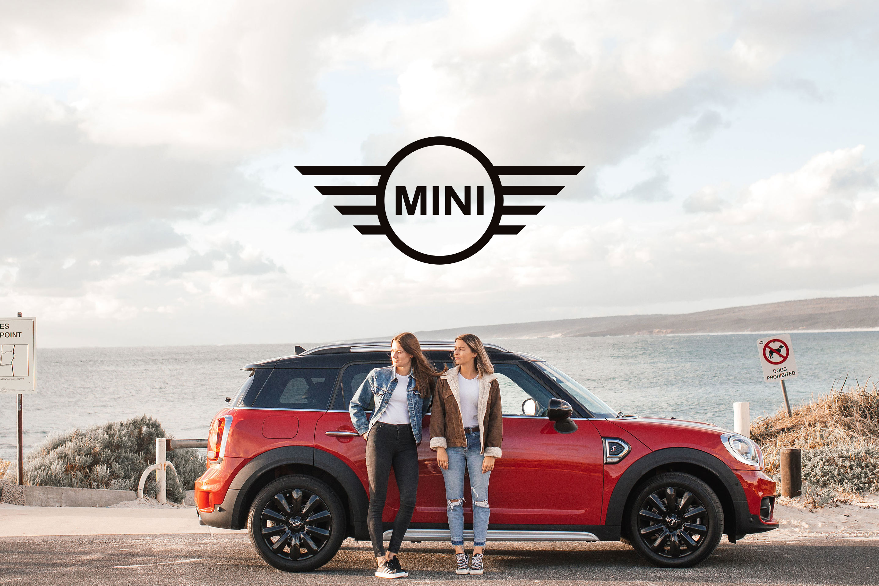 MINI : le logo de la marque dans un ciel nuageux, au-dessus de deux jeunes femmes adossées à une MINI rouge, face à la mer