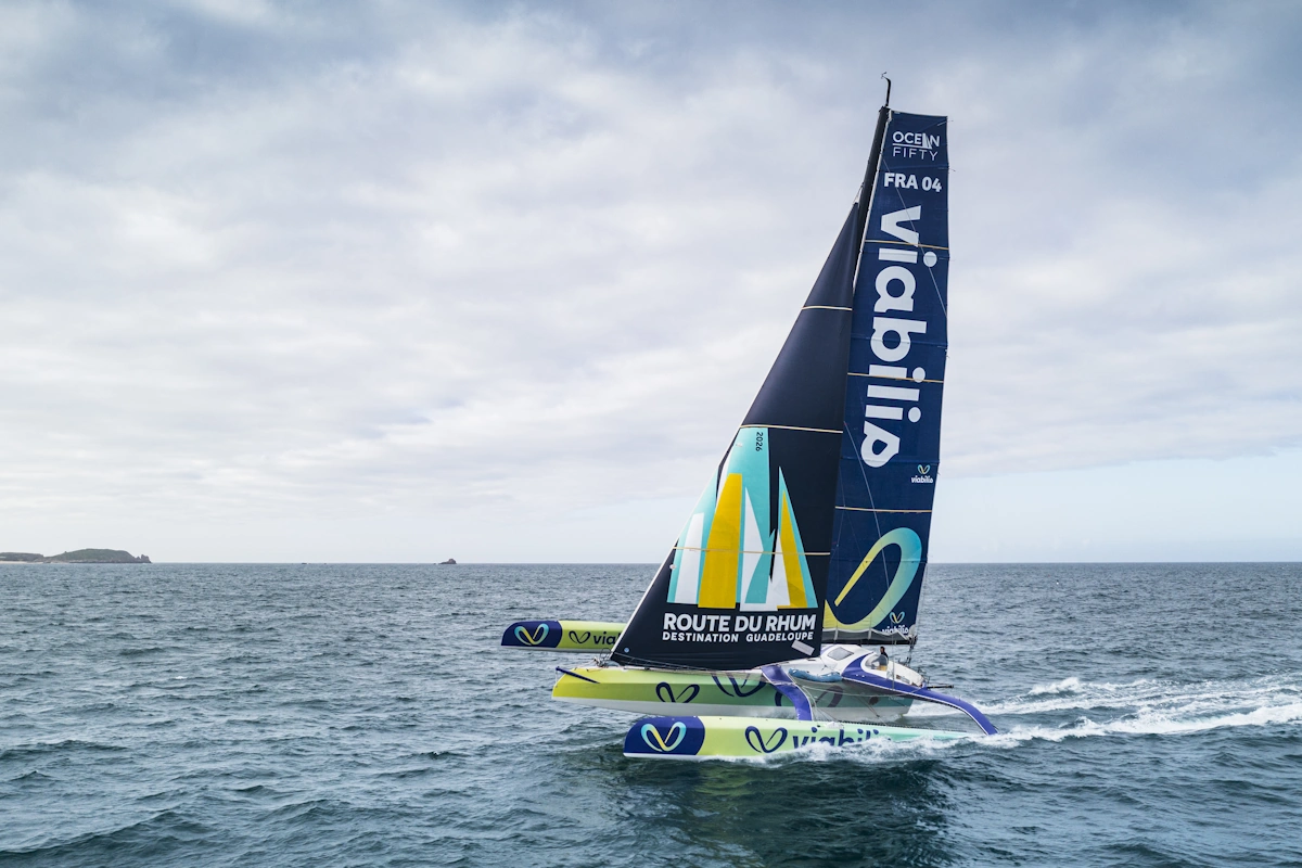 Un trimaran Ocean Fifty en mer, l’affiche de la Route du Rhum imprimée sur sa voile d’avant