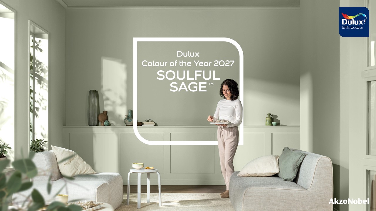Un salon peint en vert sauge clair, une femme pose un plateau près d’un canapé blanc, avec l’encadré Dulux Colour of the Year 2027 Soulful Sage