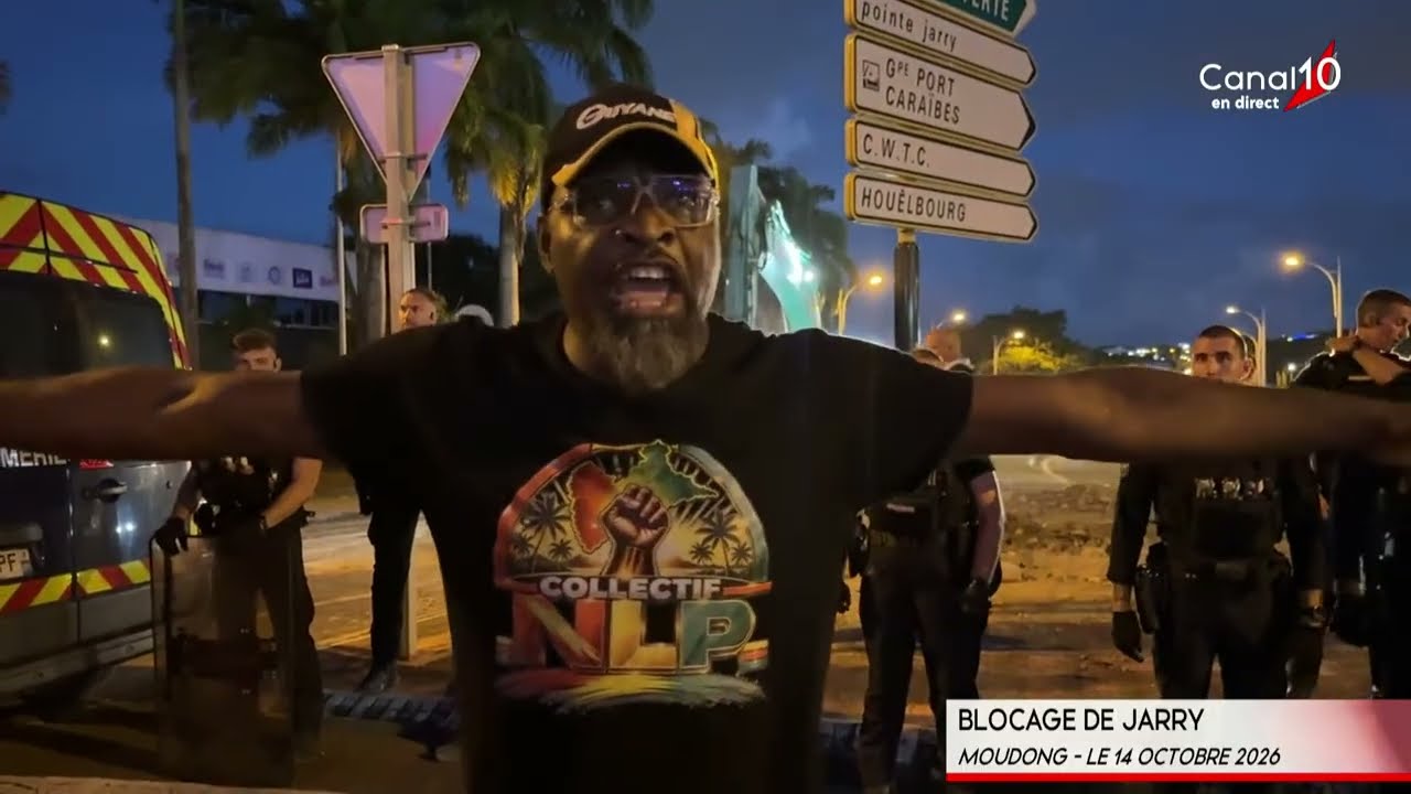 Un militant du collectif Nou Las Palé, bras écartés, devant des gendarmes, au carrefour de Moudong sous les panneaux Pointe Jarry et Grand Port Caraïbes, de nuit