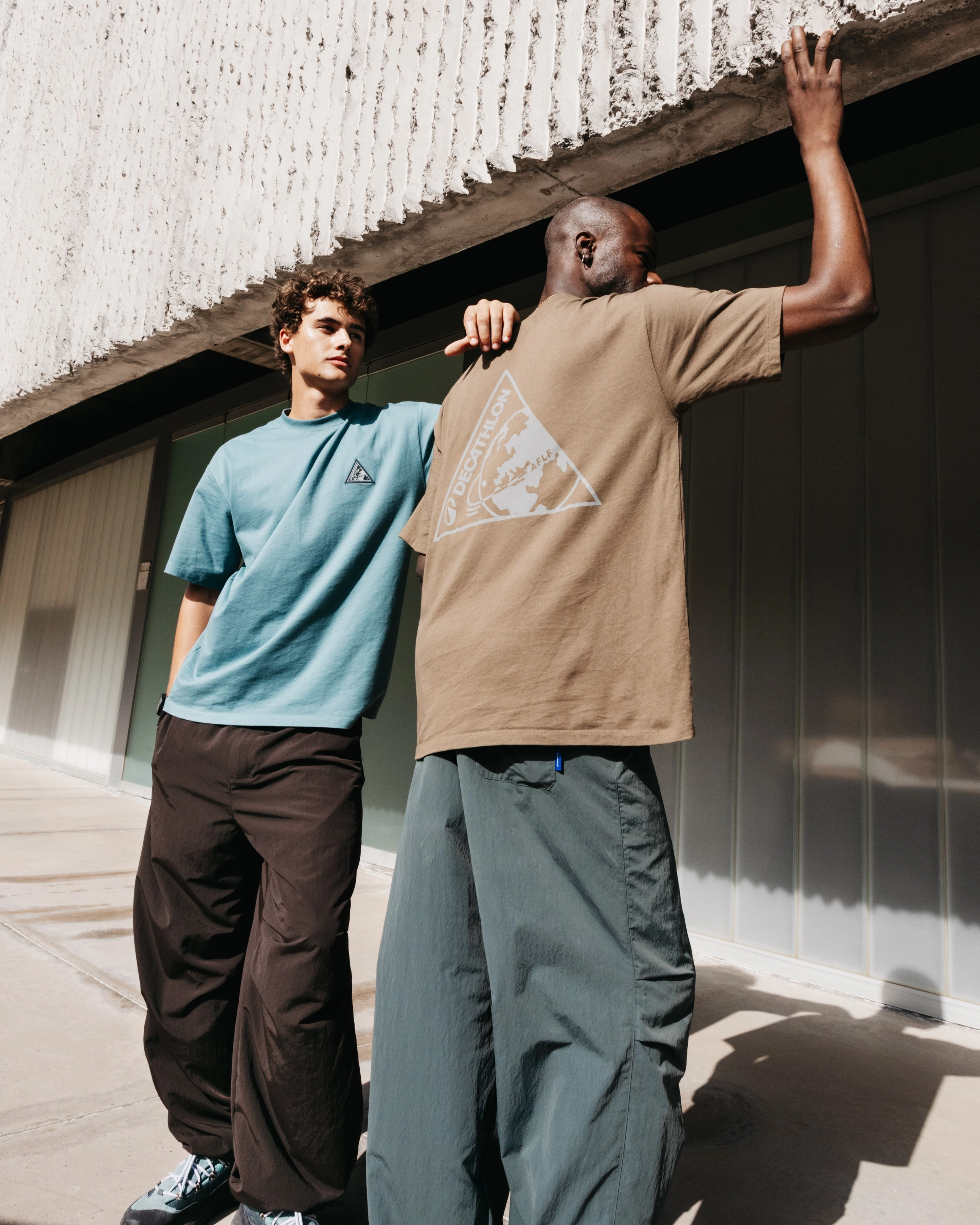 Deux hommes adossés à un mur sous un auvent de béton, l’un en tee-shirt bleu-vert, l’autre de dos, un grand triangle blanc imprimé sur son tee-shirt beige