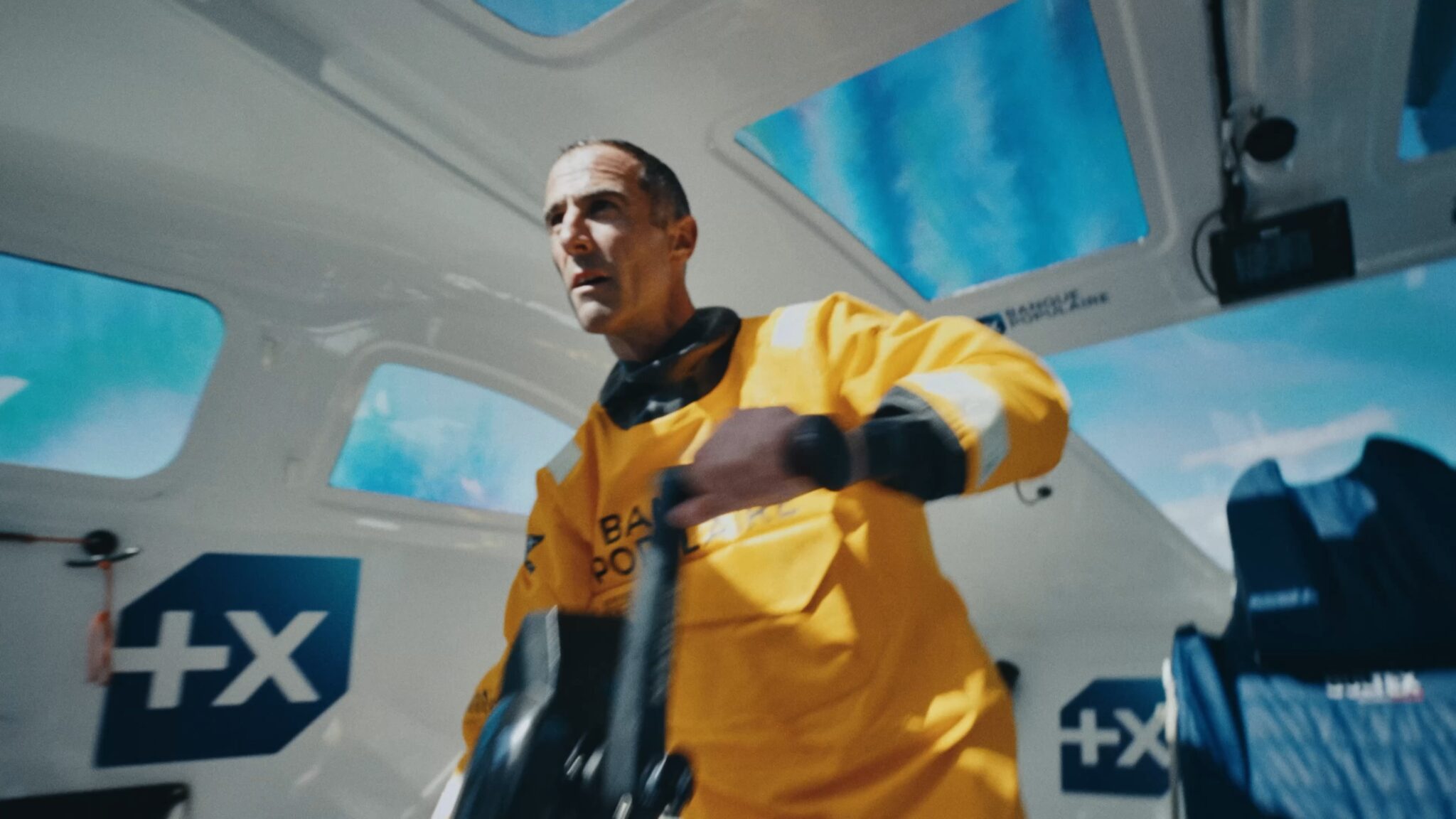 Armel Le Cléac’h en ciré jaune, à la barre dans le cockpit blanc du Maxi Banque Populaire XI, sous un ciel bleu vu à travers les hublots