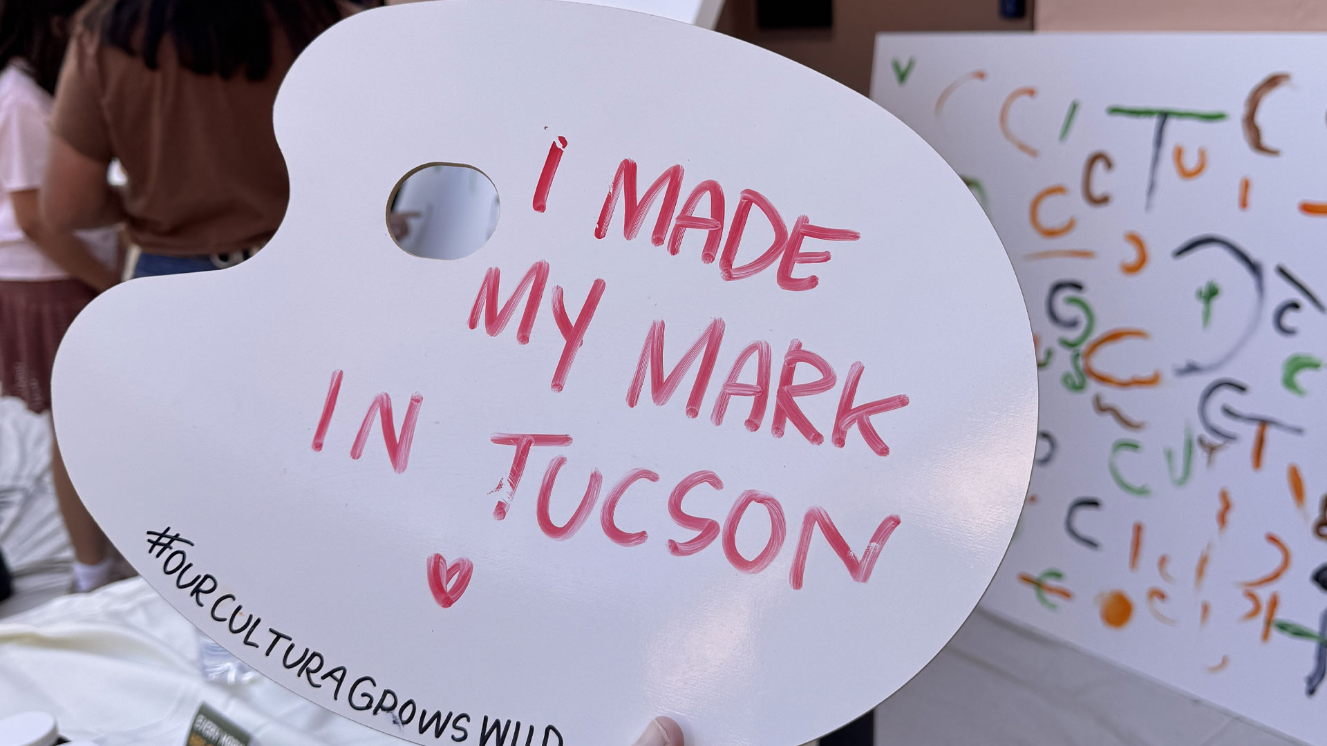 Une palette de peintre blanche où l’on a écrit au doigt, en rouge, I made my mark in Tucson, devant une toile couverte de traits et de courbes de couleur
