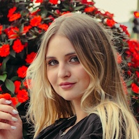 Young woman with long blonde hair smiling softly in front of red flowers.