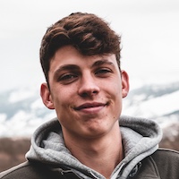 Young man with short brown hair wearing a gray hoodie, smiling, with a snowy mountain landscape behind him.