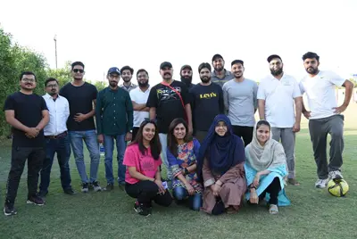 Group of 14 people posing outdoors on grass with a soccer ball, with nine men standing and five women sitting in front.