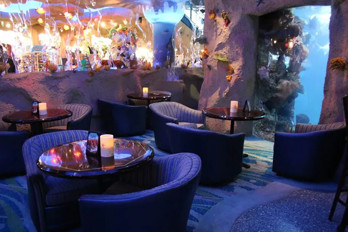 Dimly lit lounge with blue upholstered chairs and round tables, featuring underwater-themed decor and an aquarium wall.