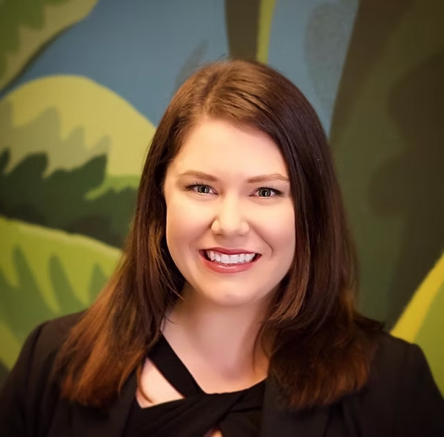 Portrait of a smiling woman with straight brown hair wearing a black blazer, in front of a colorful abstract background.