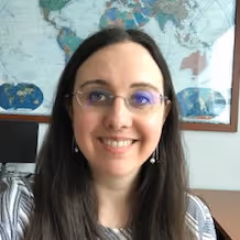 Smiling woman with long dark hair, wearing glasses and pearl earrings, in front of a world map.