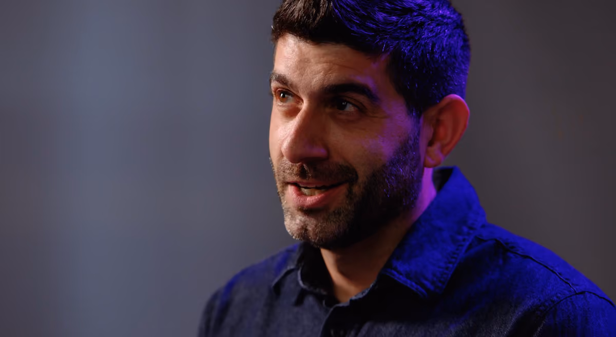 Man with short dark hair and beard wearing a dark blue shirt, looking slightly to the side with a thoughtful expression.