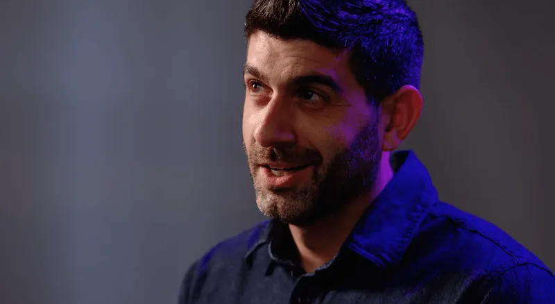 Man with short dark hair and beard wearing a dark blue shirt, looking slightly to the side with a thoughtful expression.