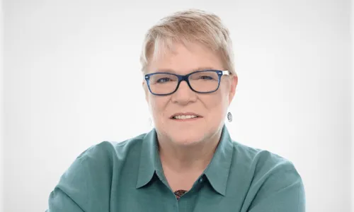 Woman with short blonde hair and blue glasses wearing a teal collared shirt against a white background.
