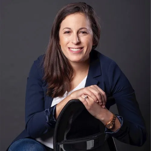 Smiling woman with long brown hair wearing a navy blazer and white shirt, leaning on the back of a black mesh chair.