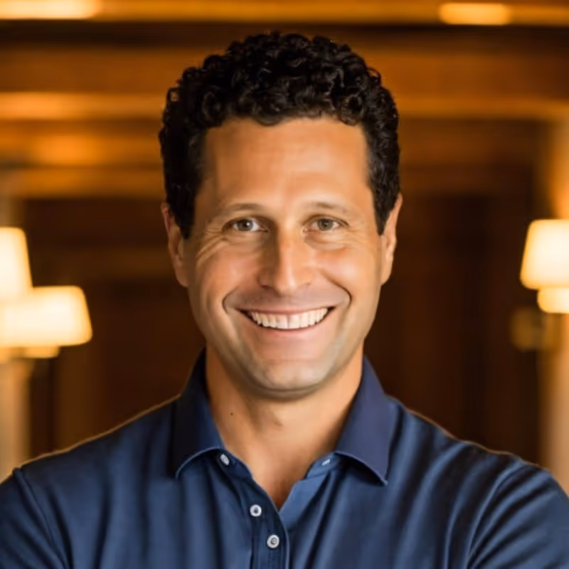 Smiling man with curly hair wearing a blue collared shirt standing indoors with warm lighting in the background.
