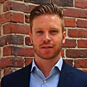 Portrait of a man with light brown hair and a trimmed beard wearing a navy blazer and light blue shirt standing in front of a red brick wall.