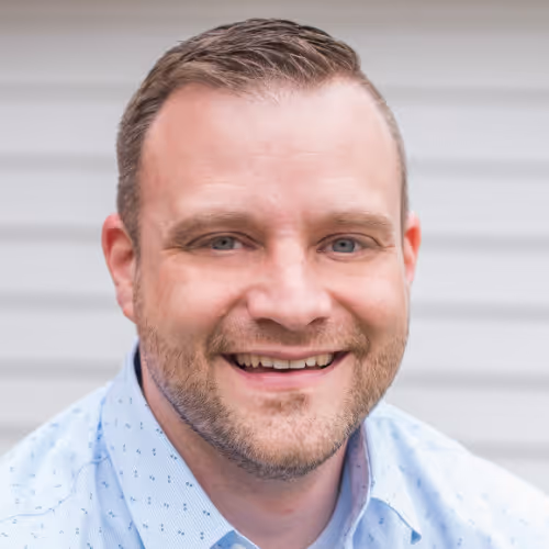 Smiling man with short hair and light beard wearing a light blue patterned shirt against a blurred gray background.