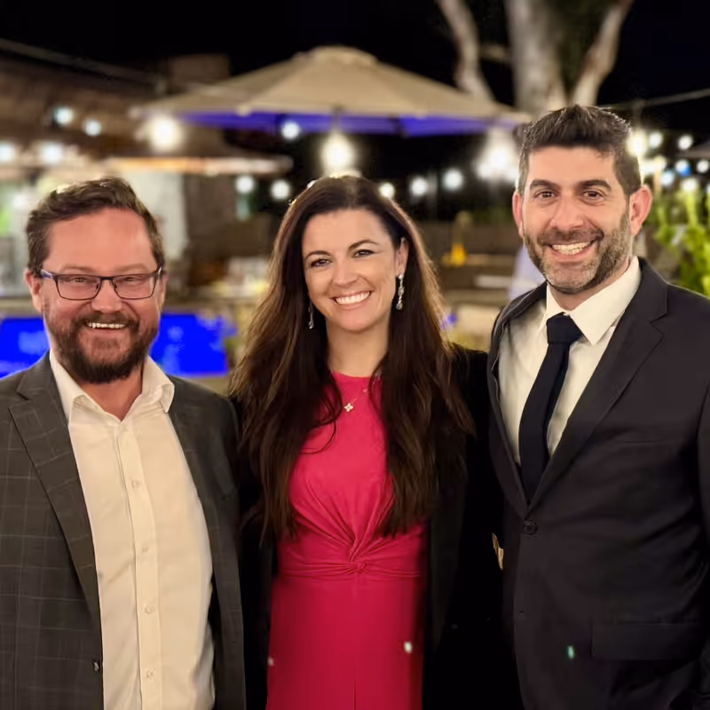 Three people dressed in formal attire smiling and standing close together outdoors at night with string lights and an umbrella in the background.