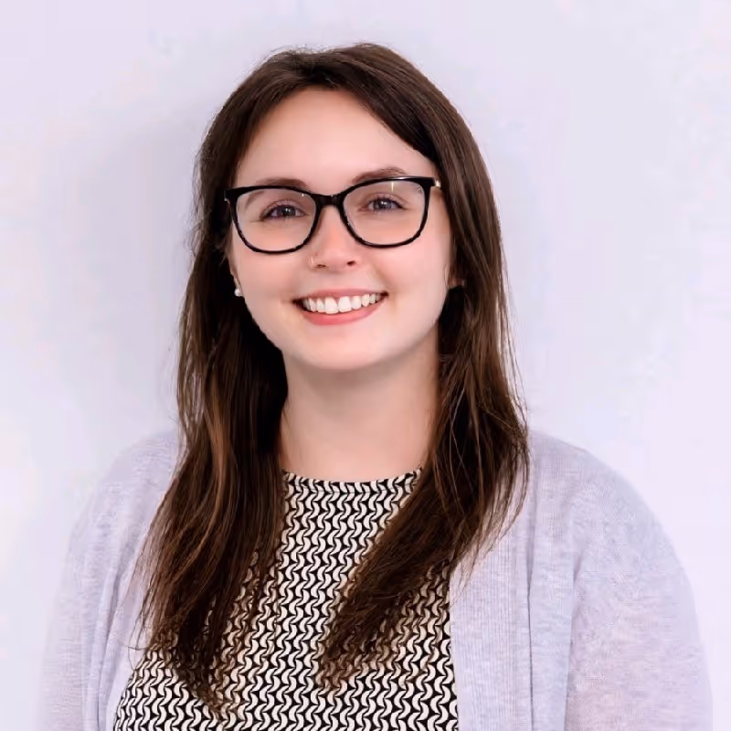 Young woman with long brown hair, black glasses, and a patterned top smiling against a light background.
