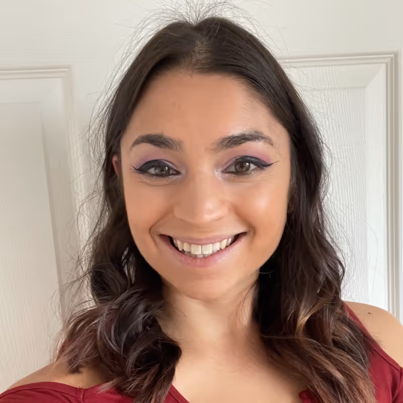 Smiling woman with long wavy brown hair wearing purple eyeshadow and a red off-shoulder top.