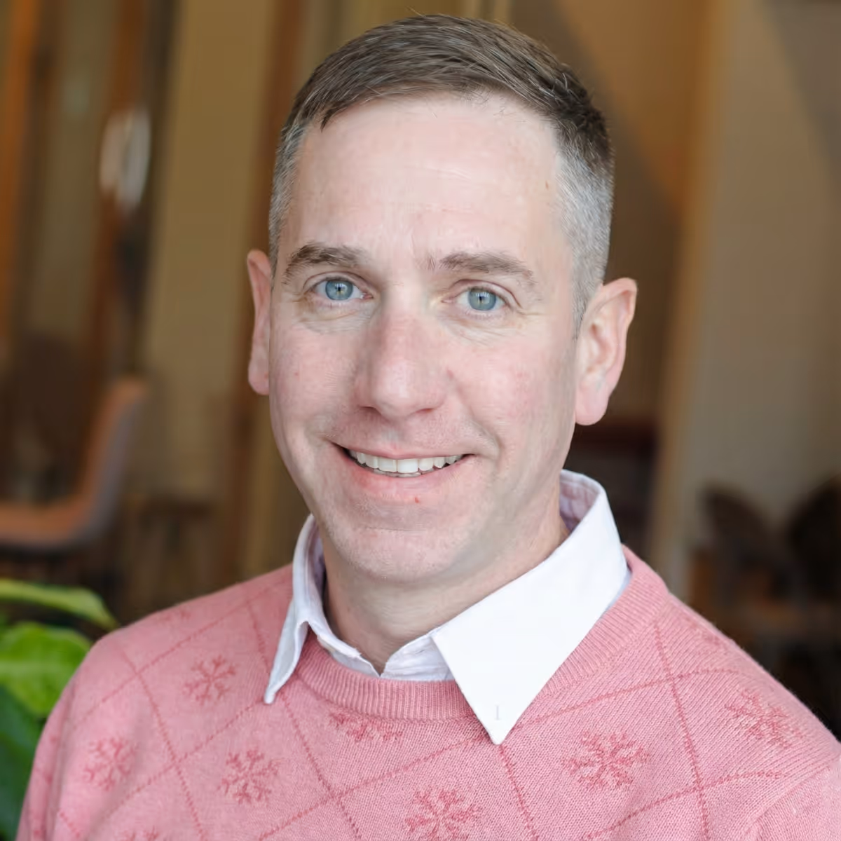 Smiling man with short hair wearing a pink sweater with snowflake patterns over a white collared shirt.