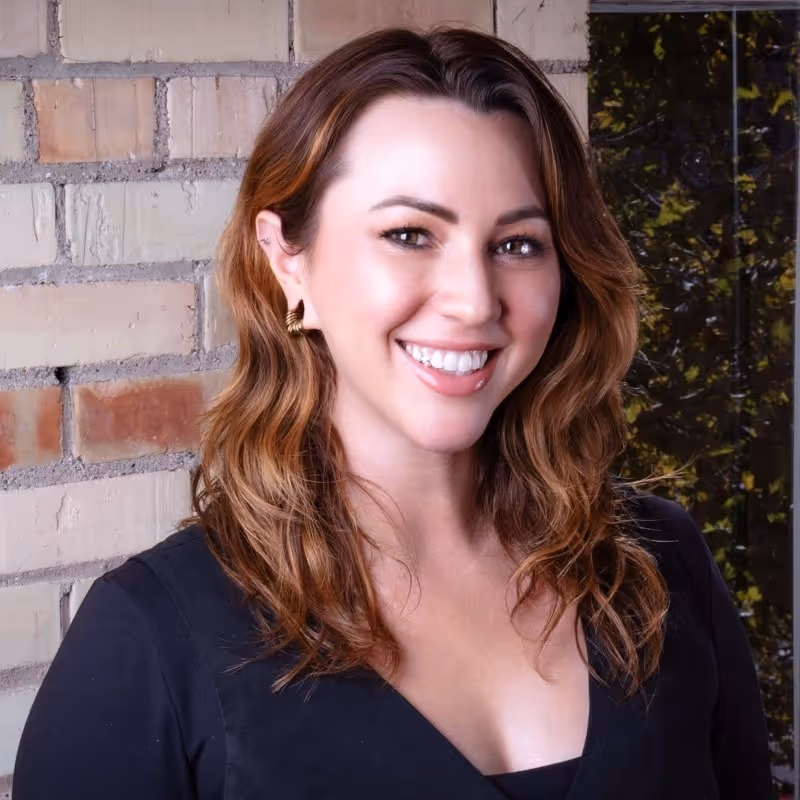 Smiling woman with wavy light brown hair wearing a black top against a brick wall background.
