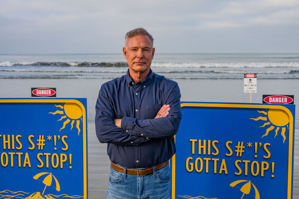 Scott Peters on a closed San Diego beach between signs reading “This s#*!’s gotta stop!” over the Tijuana River sewage crisis