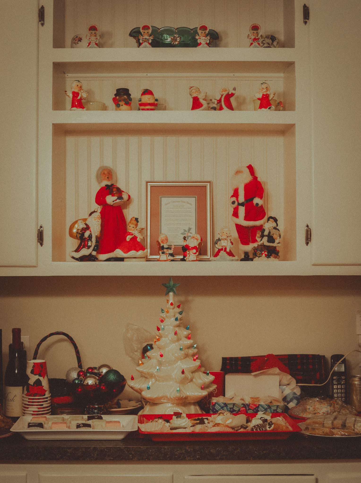 Vintage Santa and angel figurines on a kitchen shelf above a ceramic tree and trays of cookies