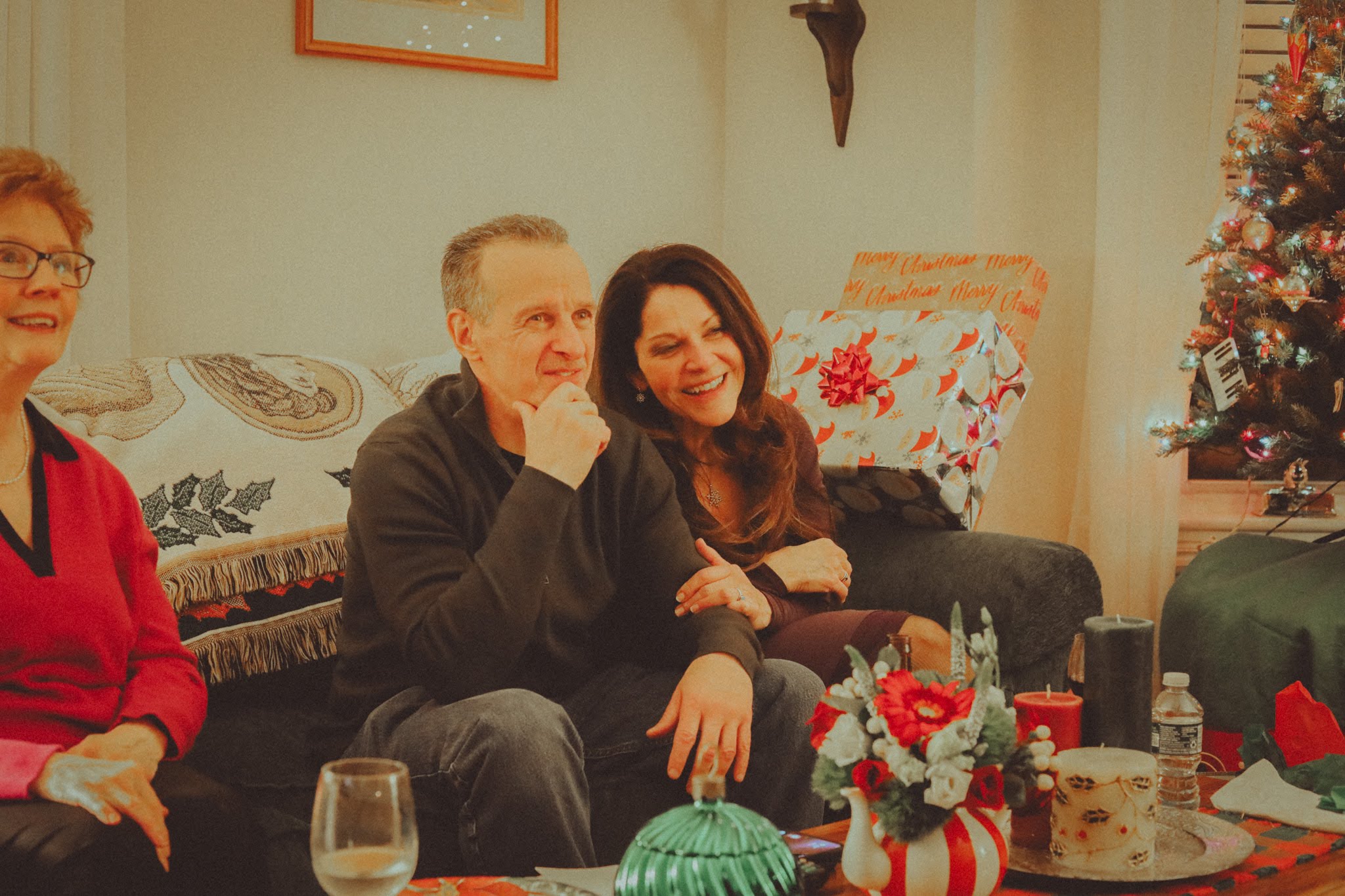 A couple laughs together on the couch beside wrapped gifts and the tree, grandma at their side