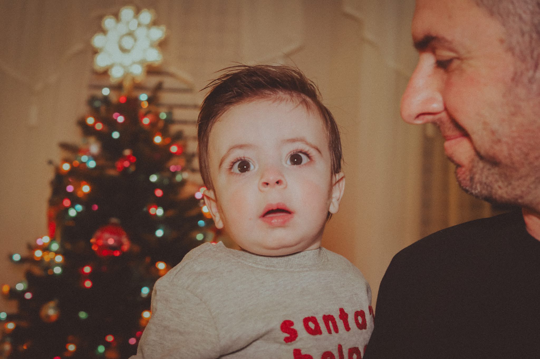 A wide-eyed baby in a Santa's helper sweatshirt, held by his dad in front of the tree