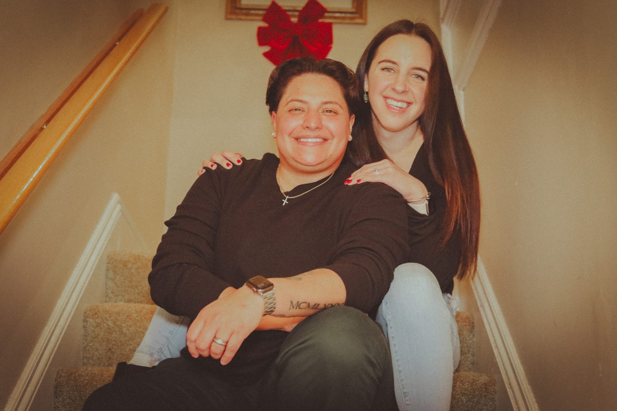 A couple sits together on their hallway stairs under a red holiday bow