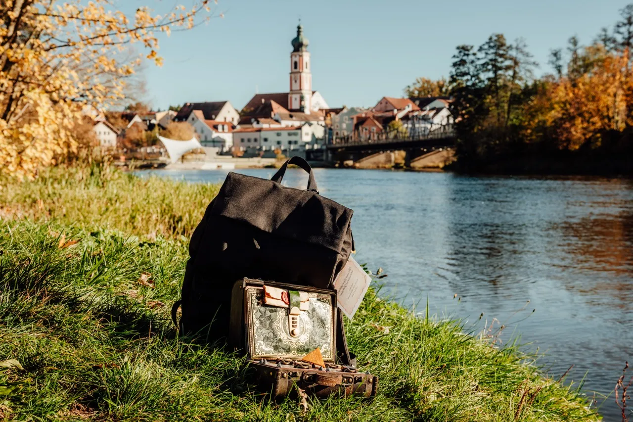 Rucksack und geöffneter Koffer im Gras am Regenufer, dahinter die Rodinger Altstadt mit Kirchturm und Brücke