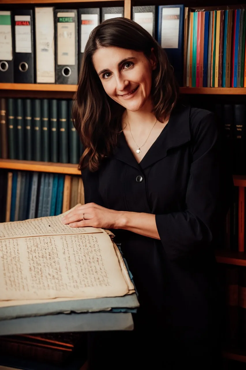 Regina Kerscher lächelnd vor einem Regal mit Aktenordnern, die Hand auf einem alten handgeschriebenen Buch