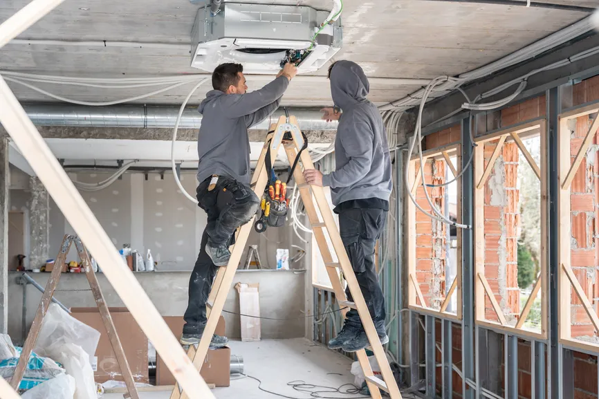 Two technicians installing a ceiling HVAC unit in a building under renovation.