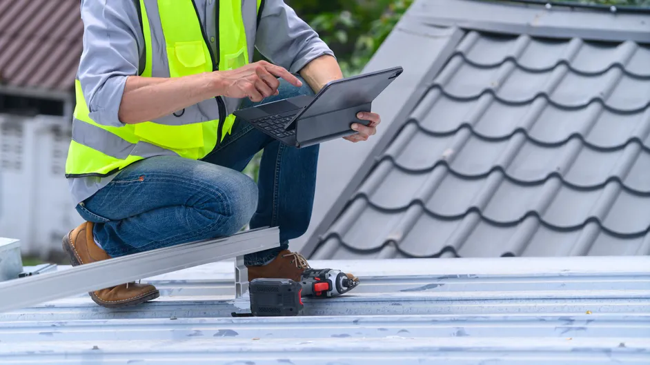 Roofer using a tablet while kneeling on a metal roof.