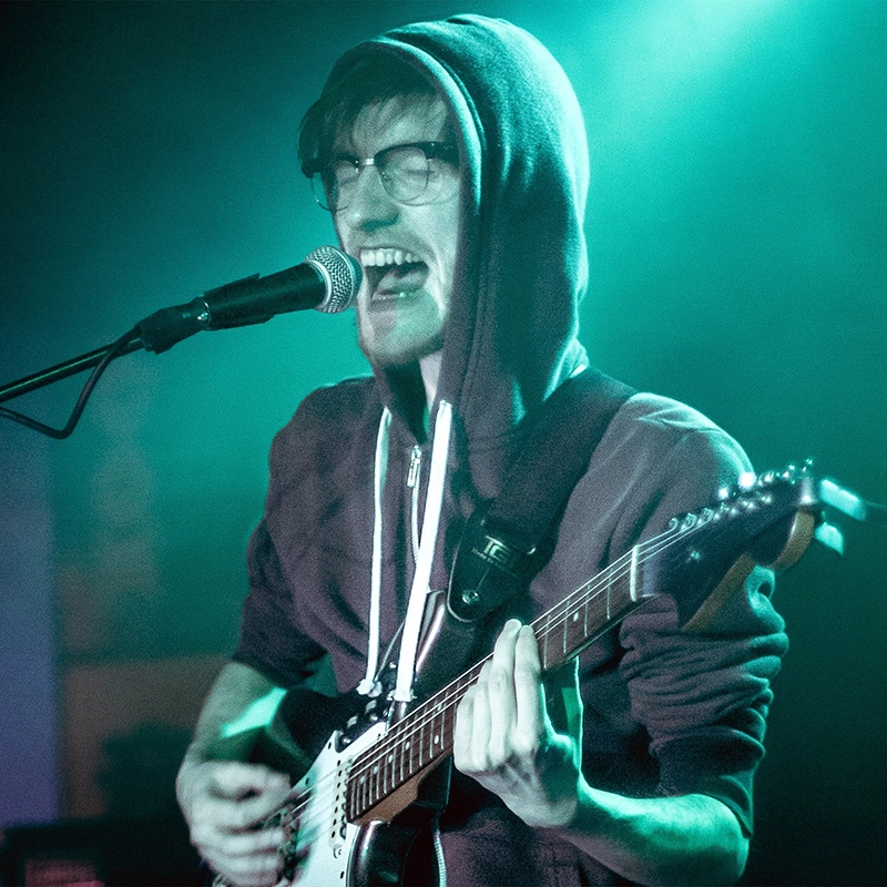 A photograph of Steve and the Sea performing live. Steve is dressed in a burgundy hoody with the hood up, and is singing loudly into the microphone whilst also playing guitar. A bright turquoise light shines from behind.