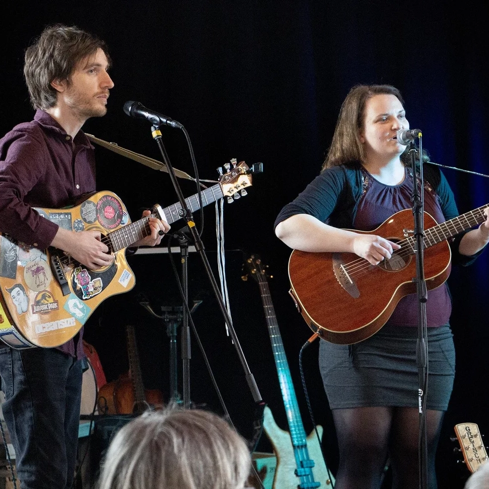 A photograph of Kerrin & McCarthy performing live. Steve McCarthy stands on the left of the image, while Vivien Kerrin stands on the right. Both are holding acoustic guitars and singing into microphones. There are various other instruments on the stage behind them.