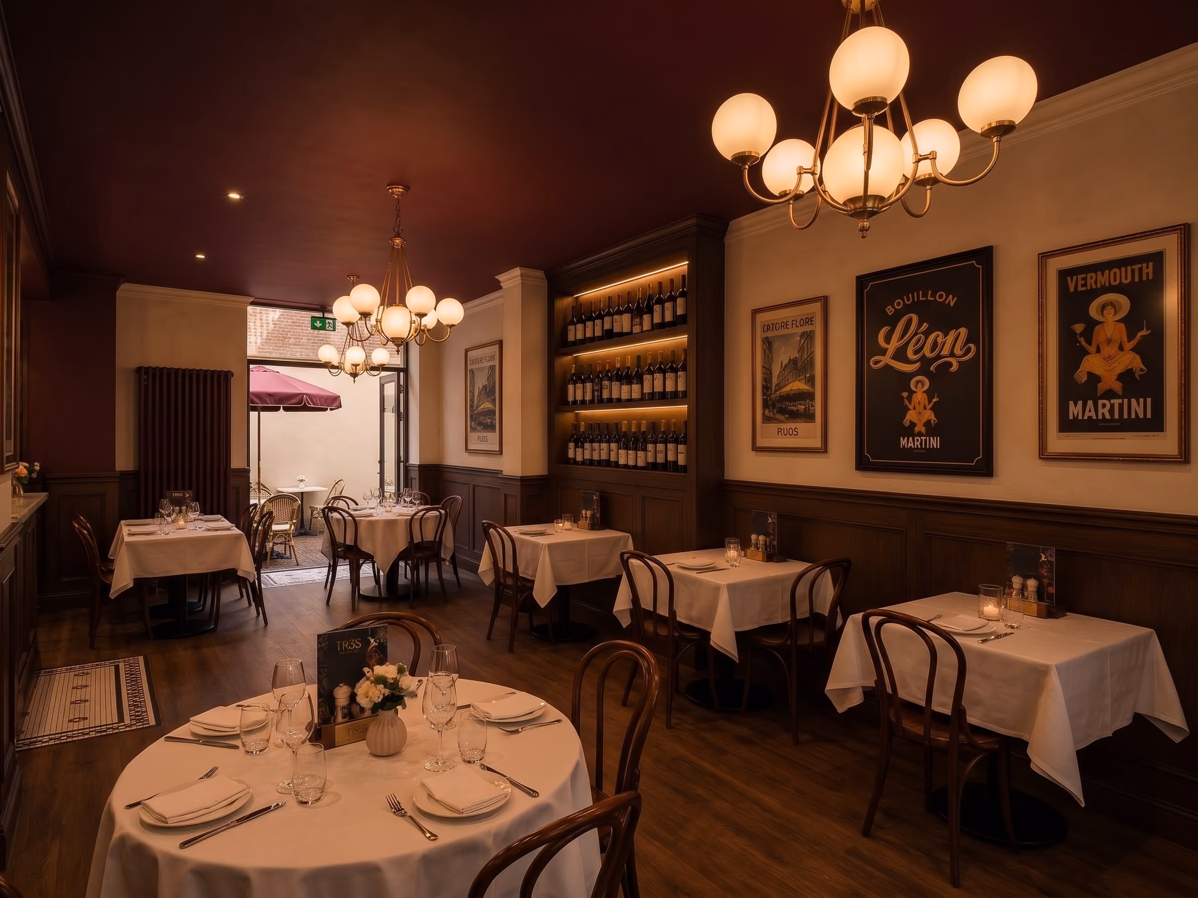 Gezellig restaurantinterieur met houten vloer, ronde en vierkante tafels met witte tafelkleden, houten stoelen, warme verlichting van bolvormige kroonluchters, ingelijste vintage posters aan de muren en een plank met wijnflessen. De achterdeur geeft toegang tot een terras met een parasol.