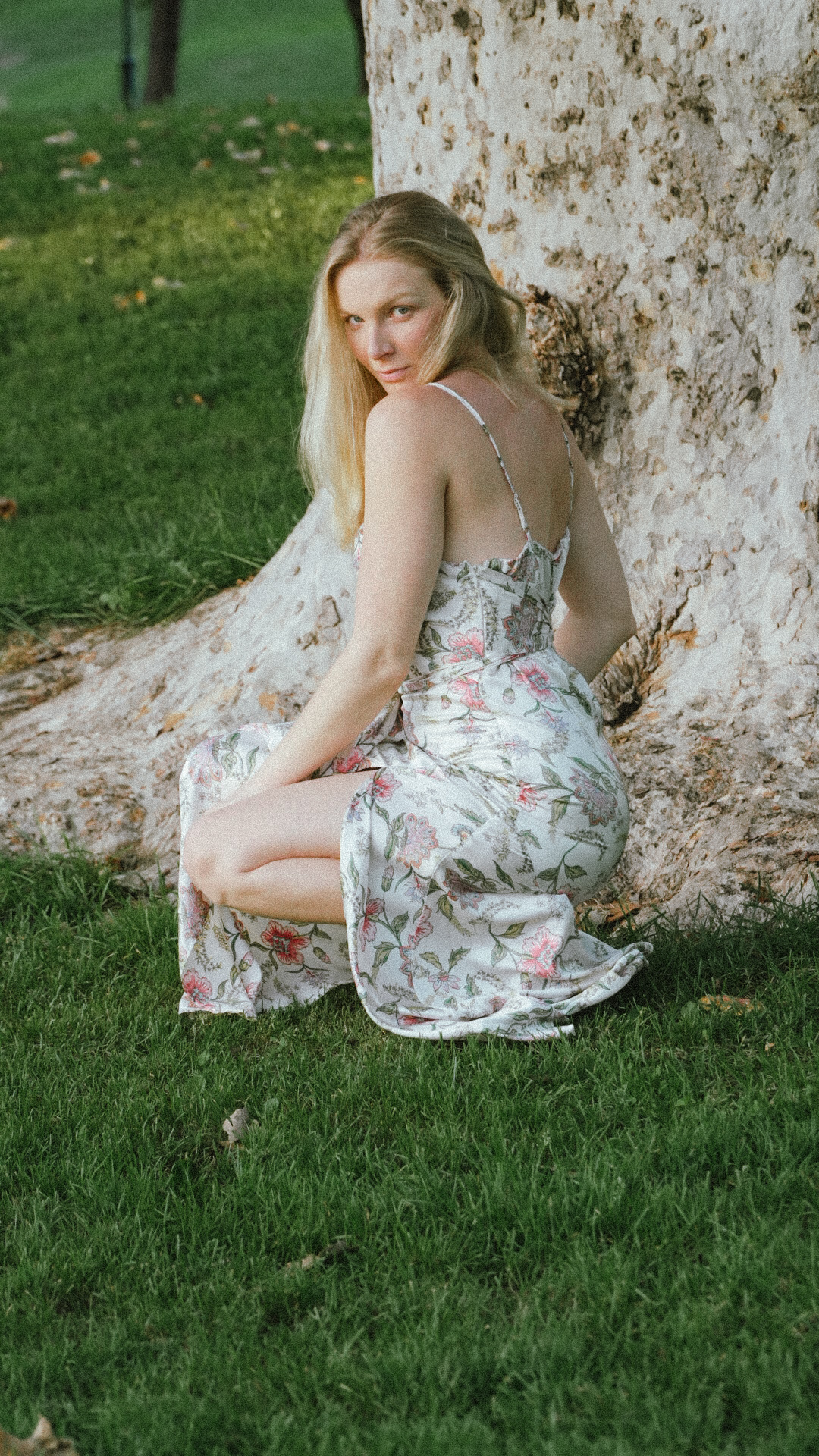 Amanda Beck in a floral dress, kneeling at the base of a large pale tree on green grass, looking over her shoulder