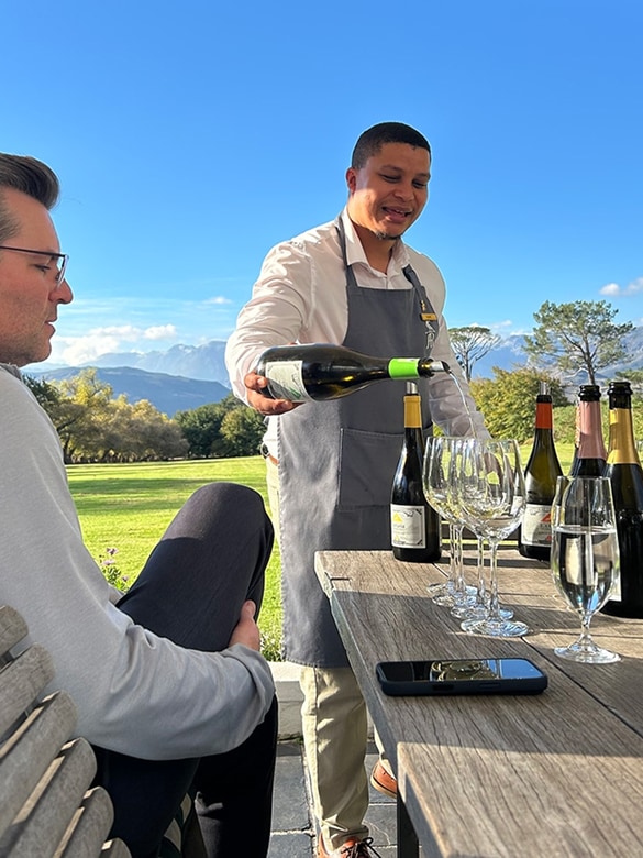 Man in a gray apron pouring white wine into empty wine glasses on a wooden table outdoors, with bottles and a phone on the table and a seated man watching under a clear blue sky with mountains and trees in the background.