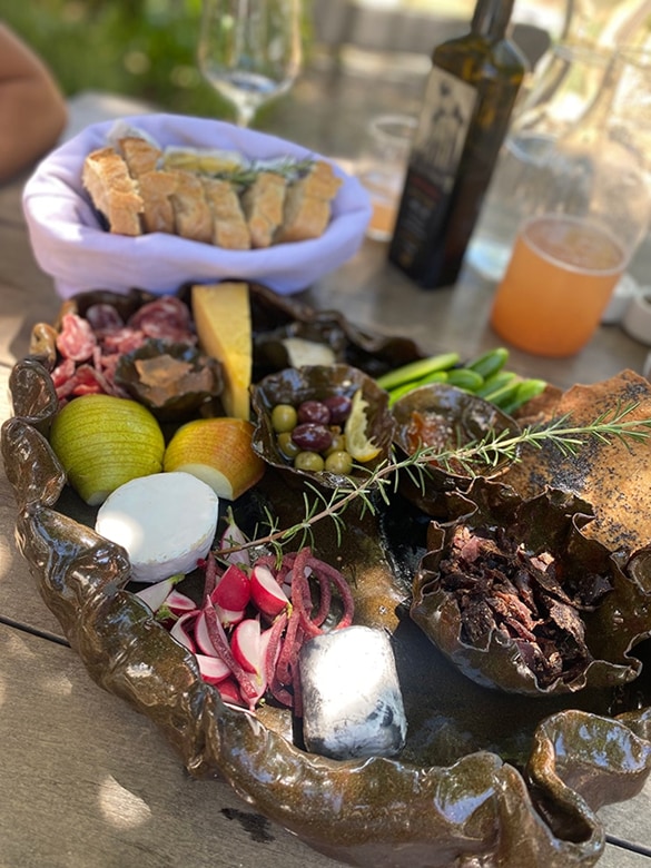 Ceramic platter with an assortment of foods including sliced bread in a cloth-lined basket, green olives, radishes, sliced pears, cheese rounds, cured meats, asparagus stalks, and thin slices of meat, garnished with a sprig of rosemary, on a wooden table with glassware and a drink in the background.