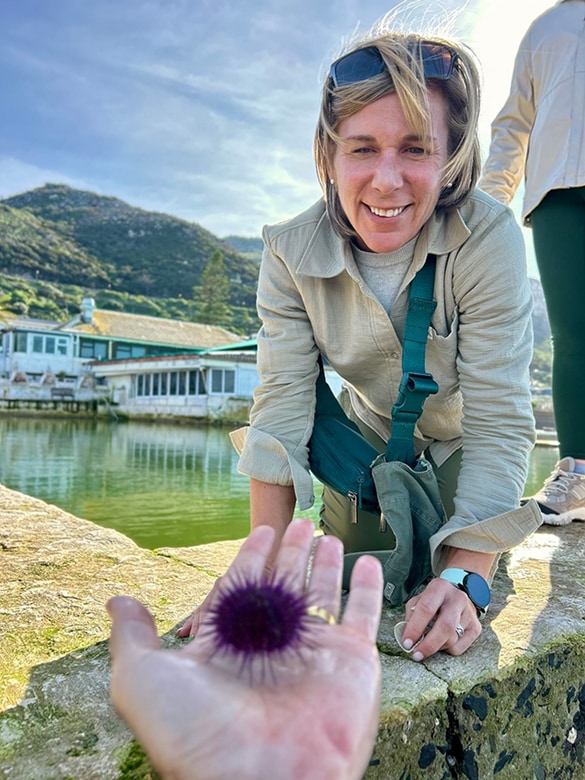 Smiling woman with blonde hair and sunglasses on her head leans forward near a body of water, looking at a person holding a purple sea urchin on their outstretched hand, with hills and buildings in the background.