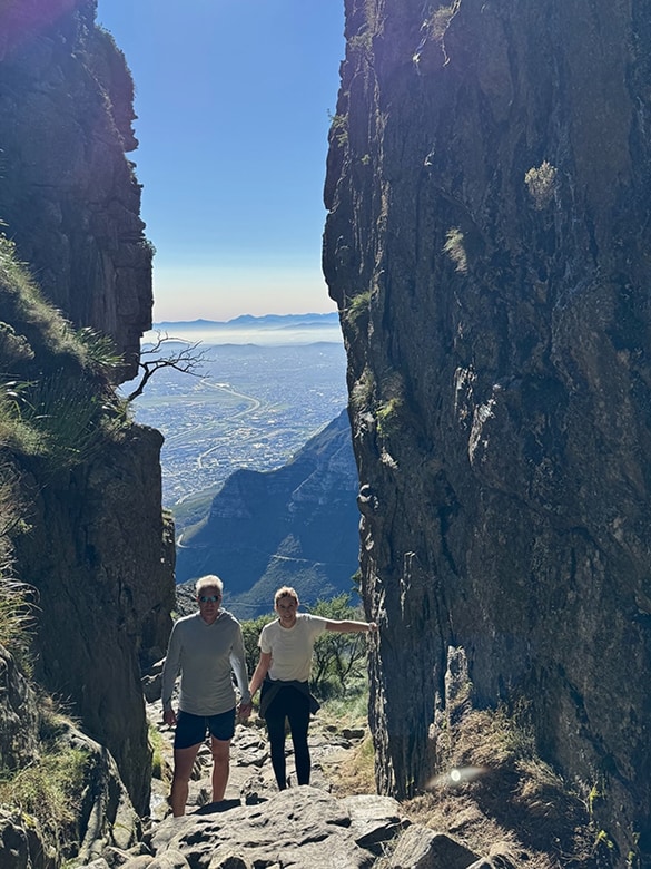 Two hikers, a man and a woman, stand on a rocky trail between tall cliffs with a spacious valley and hazy mountains in the background under a clear blue sky.