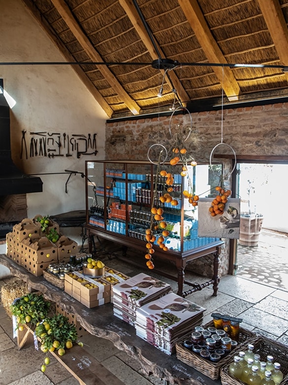 Interior of a rustic market stall with a wooden ceiling, stone walls, and a display table featuring fresh lemons, packaged goods, jars of preserves, and hanging bunches of oranges.