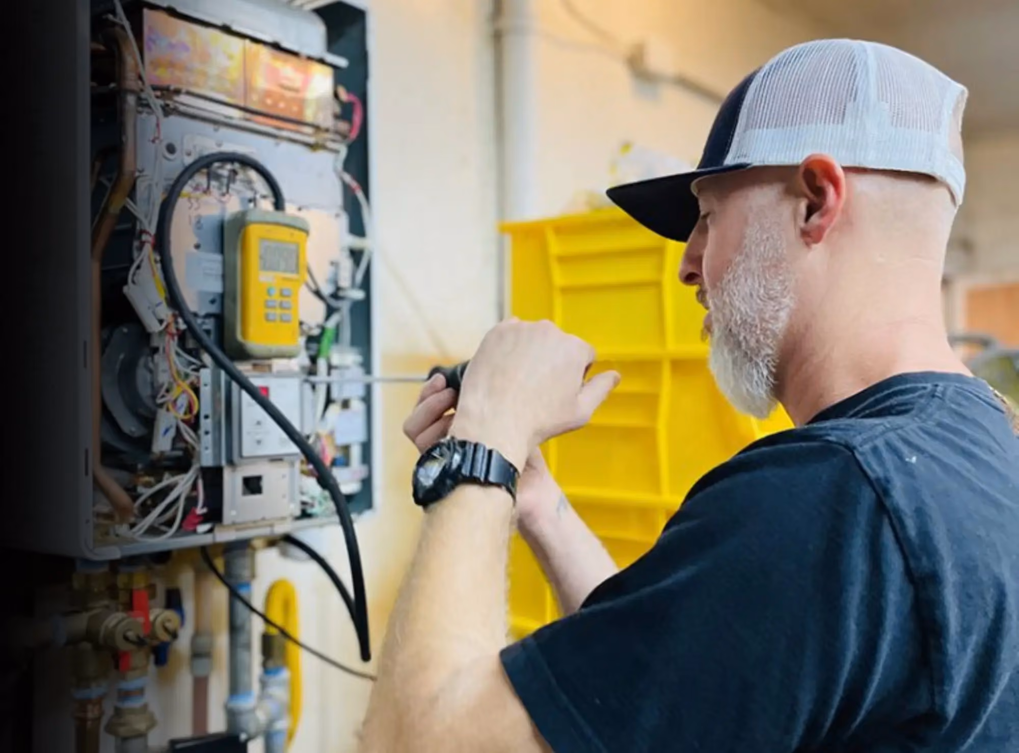 A Hot Water Guys technician in a cap and navy T-shirt works inside an open tankless water heater with a yellow digital meter clipped to the unit