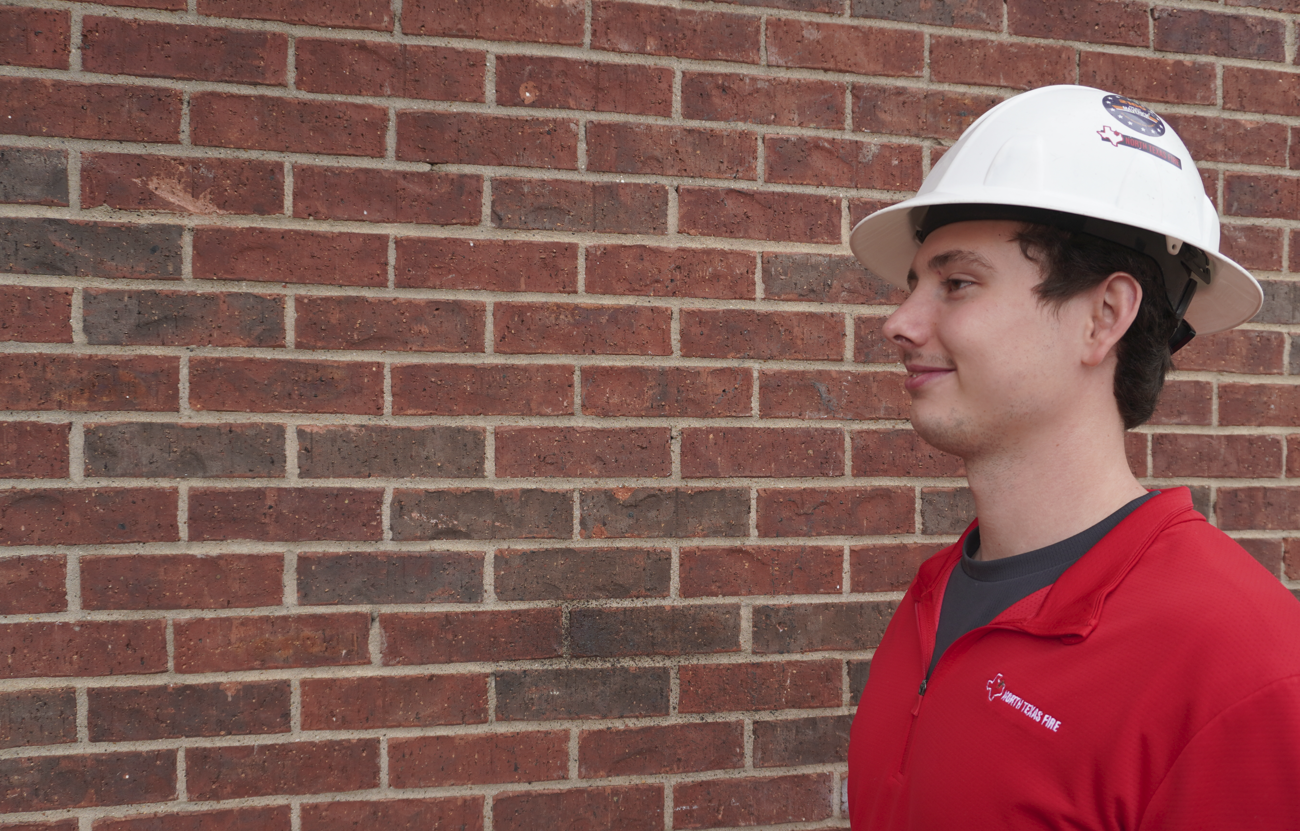 Man in red shirt and white hard hat with North Texas Fire logo stands against a brick wall.